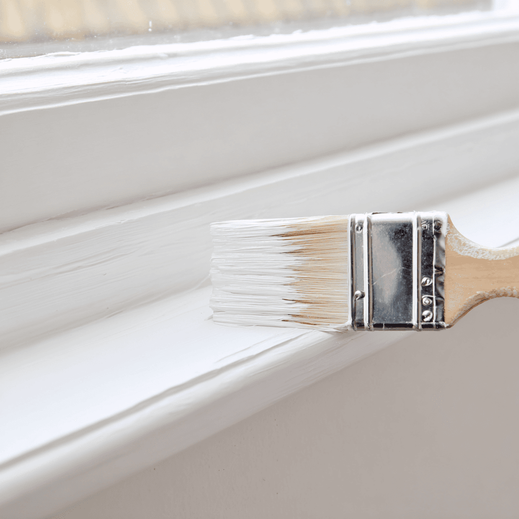 Paintbrush applying fresh white paint to window trim before property sale